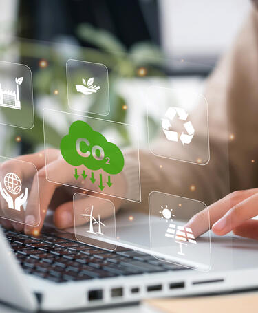 Energy_GettyImages-1401766298 Person typing on a laptop with icons hovering above the keyboard with images of hands, wind turbines, solar panels.